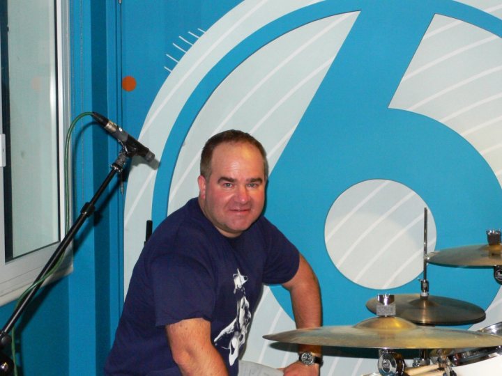 Lets  Pistonheads Gig - The image captures a moment in a studio with a predominantly blue color scheme. A man is standing behind a drum set, exuding confidence with a smile that reaches one eye. He's wearing a dark blue t-shirt and leans in towards the camera, touching the back of the chair. The drum kit and the blue backdrop suggest a focused and creative environment. The overall atmosphere is energetic and inviting, indicative of a shared passion for music.