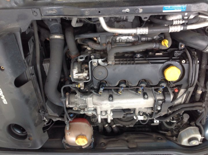 Change blow plugs - Page 1 - Saab - PistonHeads - This image shows the engine compartment of a vehicle, with various mechanical components visible. The engine has hoses and tubing systems that are interconnected, with one hose adjusting the tension on the belt. There is a capped tube laying on the engine, and a metal flange with bolts can be seen near the engine housing. The image provides a detailed look into the underhood area of the vehicle.