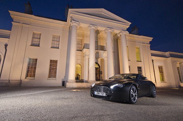 My V12V photographed by a talented photographer (not me) - Page 1 - Aston Martin - PistonHeads - The image presents a charming scene of a black convertible parked at night in front of a stately white building with classical architectural elements. The building has columns and features a brightly lit entrance. The car gleams under the night sky, adding a touch of luxury to the serene setting. The overall atmosphere is one of sophistication and quiet elegance.