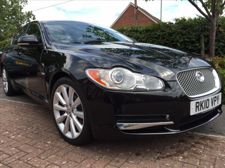 Stickering up for 2016 - Page 1 - Le Mans - PistonHeads - The image shows a sleek, black Jaguar XK convertible parked on a driveway. The car features a distinctive grille, round LED headlights, and silver accents. It is surrounded by a neatly trimmed hedge and a brick pavement. The license plate of the car reads "RK5 VPY." The uneven distribution of the shadows suggests a setting with the sun low in the sky.