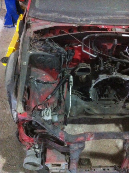 hi everyone!(expecting help)(sti 2004) - Page 1 - Subaru - PistonHeads - The image shows a damaged car front-end, situated on what appears to be a workbench or garage floor. The vehicle's front bumper is completely off, exposing the underlying metal frame and the car's electronic wiring. Visible is the hood, propped up, and a component box is open, revealing wires and hoses. The front passenger wheel is also visible, indicating that the damage extends beyond the bumper area. The damage seems severe, suggesting the car has been in a significant collision.