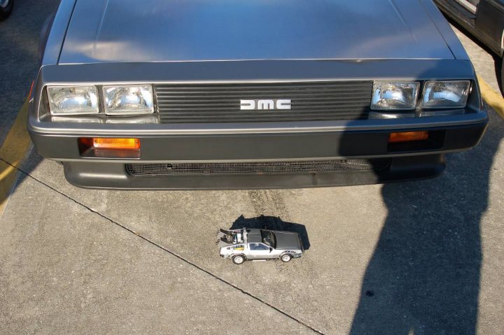 Heads Pistonheads Piston Delorean - In the image, a small, silver toy car is seen prominently in the foreground on a road, facing away from the camera. Behind it, a blue AMG car is parked with its headlights on, and a man is on a pedestrian crossing, partially visible and casting a shadow. The road appears wet, and there's a yellow line visible on the concrete. The juxtaposition of the toy car in front of the larger vehicle creates a contrast from the two different scales of objects in the frame.