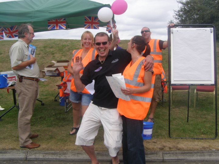 Growl Pistonheads Griff - In the image, a group of people is engaged in a lively outdoor gathering. At the center of the group, a tanned man dressed in a black shirt and orange safety vest is balloon dancing, creating a joyful atmosphere. A blonde woman, wearing a bright orange vest, is standing next to him, laughing and enjoying the moment. Surrounding them, other individuals, some donning safety vests as well, are watching the spectacle with joy. A crimped canopy provides shade, and a deck of cards can be seen among the items on the lush grass, hinting at leisurely activities.