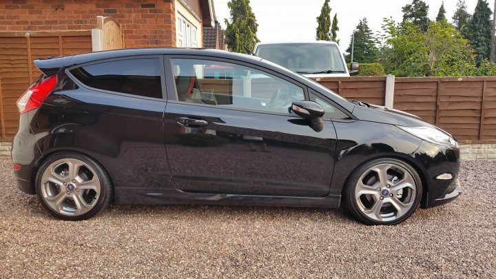 Fiesta ST Motorway Hack! - Page 2 - Readers' Cars - PistonHeads - The image shows a black car parked on a gravel driveway. The car is positioned facing the viewer with the front of the vehicle in clear view. Its wheels are silver, and it features black rims. The car appears to be a hatchback with a visible antenna on the roof. Behind the car, a residential house can be glimpsed, along with a wood fence. The sky is visible in the background, suggesting that the photo was taken during the day under overcast or cloudy conditions.