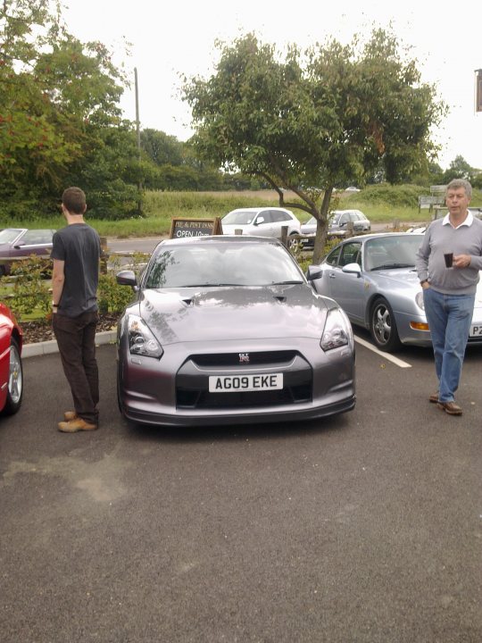 Pistonheads - The image captures a serene outdoor scene featuring a gray sports car and a silver sedan parked in a lot. A man stands by the edge of the concrete, separated from the cars by a small barrier. Another person is seen walking across the lot, past the sports car. The setting is calm, with backgrounds of trees and a second car. The scene conveys a tranquil atmosphere, possibly during a visit to a car show or a car scene in a park or open space.