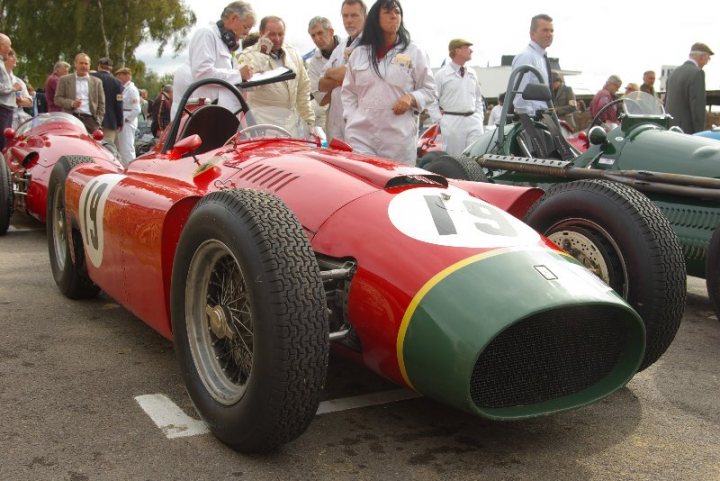 Revival - First Time and Excited - Page 1 - Goodwood Events - PistonHeads - The image captures an excited scene at a race car exhibit. A prominent red race car, bearing the number 15 and marked with a downward-pointing arrow on the hood, takes center stage. The car's open front bonnet reveals the pistons and engine, hinting at the powerful machinery within. Surrounded by people in various attires, the car appears to attract considerable attention. In the background, additional vintage racing cars, each marked with individual numbers and logos, can be seen, providing a fascinating glimpse into the rich history of racing cars.