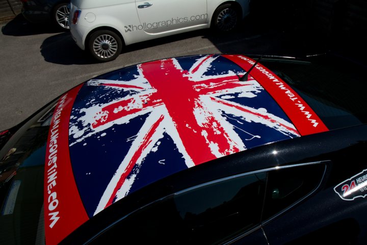 Stickered Pistonheads - The image showcases a vehicle with a unique body art featuring a British flag design on its rear bumper. The flag is prominent and takes up a significant portion of the image. The design has a slightly distressed pattern, adding texture and depth to the overall look. The vehicle is white and parked in a lot, and the flag's bold colors contrast sharply against the car's body. The image has a gritty feel, suggesting it might have been taken in an urban or industrial area.