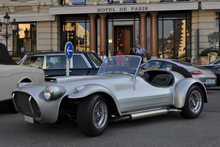 Ferrari Panther Pistonheads - The image depicts a silver, vintage-style sports car in the middle of the streets of Paris, France. The car is positioned in front of a building with a sign that reads "HOTEL DE PARIS." The car's convertible top is down, allowing the driver to enjoy the surroundings. There are people standing next to the car on the sidewalk, and the background is filled with classic cars lined up on the street, adding to the ambiance of the city. The day appears to be fair, with people and cars moving about as one would expect on a typical day in a bustling city.