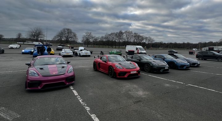 Pistonheads - The image shows a parking lot filled with various high-performance cars. There are multiple sports cars in the lot, including a bright red one and several others that appear to be luxury or supercars. They are parked side by side, demonstrating their impressive size and design. In the background, there is a skyline with trees, suggesting an urban environment. The lighting suggests it might be late afternoon or early evening.