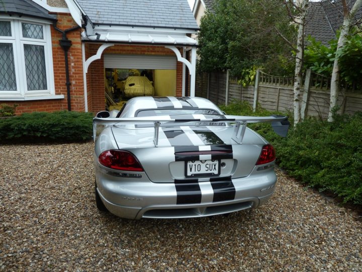 A white car parked in front of a house - Pistonheads - The image depicts a close-up view of a parked silver sports car featuring a distinctive black and white striped roof, commonly referred to as a "roof bomb" style. The car is parked on a gravel driveway next to a residential house with a garage door open. The license plate is visible and reads "V10 SUX." There are mature trees and shrubbery visible in the background, indicating a suburban setting. The overall style of the image is casual and documentary, capturing a moment in a home setting.