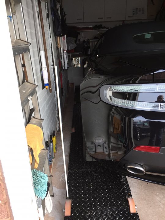 Narrow Garage - Possible Solution?  - Page 1 - Aston Martin - PistonHeads - The image shows an indoor setting, most likely a garage or storage area. A black car is parked inside, taking up the majority of the space. To the right, there's a set of shelves with various objects on them, and above the car, there's a shelf holding more items. On the left side, there's a vehicle hoist, and below it, a red carpet runs along the wall. The floor appears to be made of concrete, and a white garage door is visible in the background, providing access to the outside world.