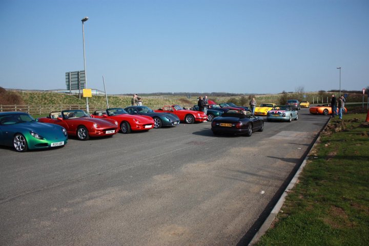 Pistonheads Newby Convoy Hall - The image showcases a diverse collection of sports cars parked in a lot on a sunny day. The cars range in color and model, indicating a gathering or event dedicated to these luxury vehicles. Spectators are scattered around the perimeter of the display, enjoying the array of vehicles. The cars are positioned on an asphalt lot with a grassy area visible in the background. In the distance, there's a glimpse of a highway on-ramp.