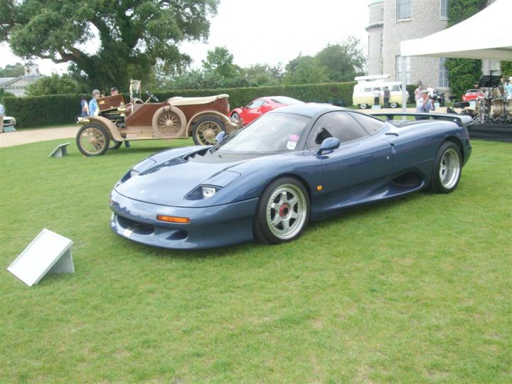 GRRC Open Day - 11 August - Page 1 - Goodwood Events - PistonHeads - The image displays a scene at a classic car show. A blue sports car, characterized by its sleek, low-profile design, is prominently parked on the grass, facing towards the left side of the photo. Behind the sports car, an array of older-model cars is arranged in a line, exhibiting a variety of colors and designs. In the background, beyond the line of cars, there is a crowd of people milling about, seemingly enjoying the event. The setting appears to be outdoors, likely a fairground or similar venue, with the cars arranged on a grassy surface.