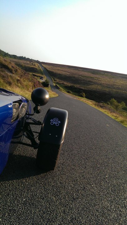 Lion inn , blakey ridge - Page 1 - North East - PistonHeads - The image shows a paved road viewed from the perspective of a vehicle traveling down the path. The road curves in the distance and is surrounded by a large, open field on both sides. A blue car is partially visible in the foreground, with a black backseat carrier attached to it. The sky is clear, suggesting it is a sunny day, and there is a logo or emblem on the black carrier. The overall scene gives a sense of solitude in a vast, open space.