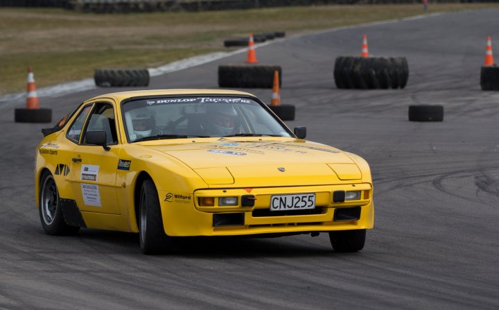 Targa 2013 - Page 1 - New Zealand - PistonHeads - The image portrays an aftermarket yellow racing Puma car on a race track. This car is equipped with various performance upgrades, such as a carbon fiber hood, a windshield with a name tag, and spoilers. It's maneuvering around a course with multiple tires raised on safety cones, suggesting active use of the track for testing or practice. The style of the image is dynamic, capturing the car in motion and the surrounding environment with motion blur.