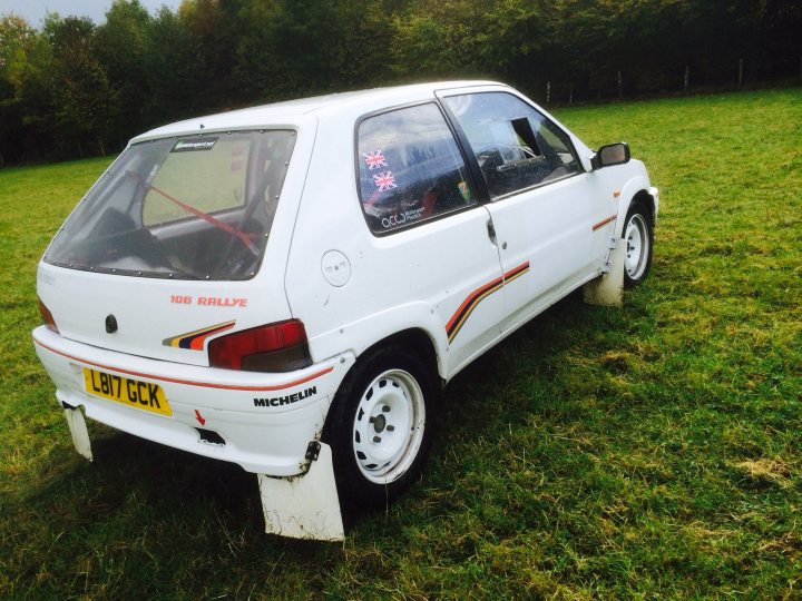 Stage Rallying - the costs? - Page 1 - UK Club Motorsport - PistonHeads - The image showcases a white car parked on a grassy field during the daytime. The vehicle appears to be relatively old, with a visible dent on the front passenger side door. There are distinct graphics and red stripes on the hood of the car. The side of the car has the text "MAX 198 MICHELIN" and a phone number which suggests it may be an advertisement or promotional vehicle. The car's registration plate reads "V6 REM". The setting suggests a countryside or suburban location, with other lush green grass around the car and trees visible in background.