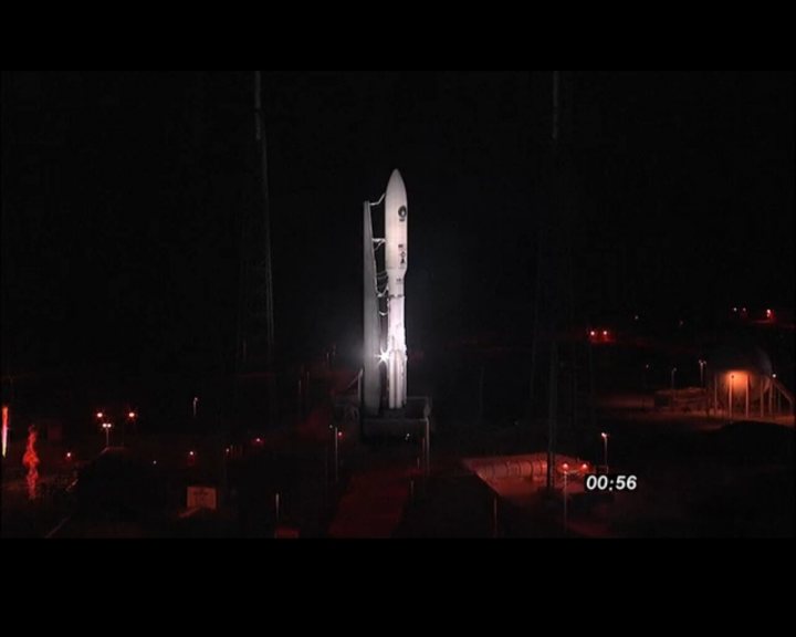 Atlas V launch tonight - Page 1 - Science! - PistonHeads - This image captures a night scene viewed from a distance, featuring a prominent rocket launch vehicle. The rocket is mounted on a metallic structure, illuminated against the dark sky. The metal structure supports the rocket and is surrounded by various lights, likely from the launch facilities or satellite uplink equipment. Beneath the launch vehicle, there appears to be a concrete platform or walkway. In the bottom right corner, a timer indicates the launch time as 00:56, which is likely the schedule for the impending rocket launch. The image conveys a sense of anticipation and the prestige of space exploration.