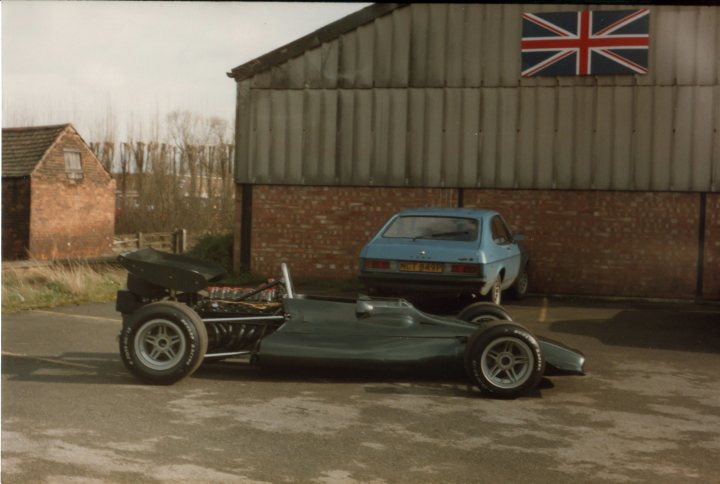 F1 from yesteryear - when F1 wasn't just about money Bernie! - Page 1 - General Motorsport - PistonHeads - This image features a green, vintage-style low-drag or go-cart combined with a body that resembles that of a F1 race car. The vehicle appears to have a black frame and it is parked next to a short, blue car. In the background, there is a partial view of a building with a large British flag flying from it. The style of the image suggests a retro or historical context, possibly a photo from several decades ago captured during the day.