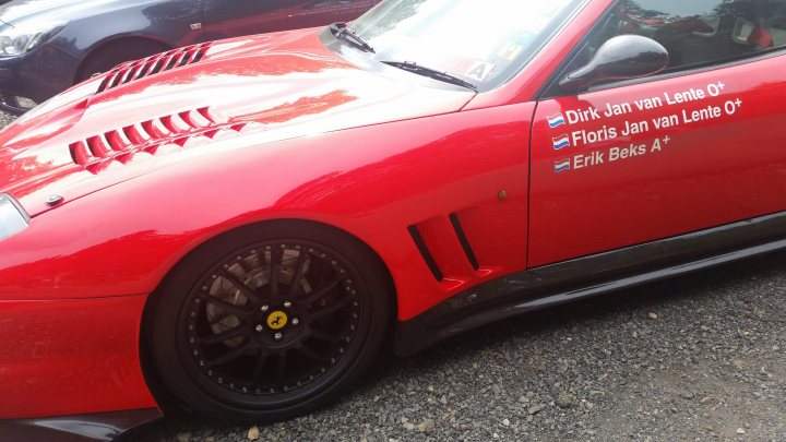 Ferrari 488 GTB at Spa F1 last weekend - Page 1 - Ferrari V8 - PistonHeads - The image shows a red sports car parked on a gravel surface. The car is angled towards the viewer, allowing for a clear view of its design and details. A notable feature is the vehicle's number, which is written in German on its side. Additionally, the car has a yellow emblem adhered to its hood, indicating that it may be a racing or track car. The background of the image is slightly blurred but suggests that the car is parked on a street with other cars, providing a sense of context and location.