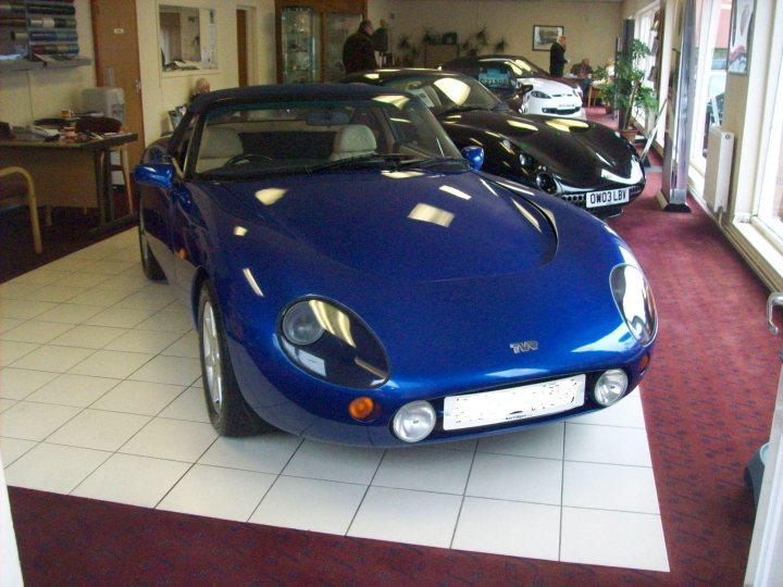 Shes Finally Pistonheads - The image features a striking blue sports car situated on a shiny, white tiled floor. The car, with its vibrant color and sleek design, is the focal point of the scene, drawing attention with its central position. The background is a tasteful blend of neutral tones, providing a contrast that further emphasizes the car's presence. There are several other cars neatly parked in the background, suggesting this might be a showroom or a special events space.