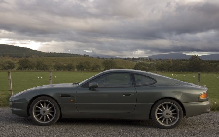 Your DB7 pictures. - Page 1 - Aston Martin - PistonHeads - The image depicts a dark gray or purple sports car on a gravel road. The car is positioned in the foreground and is the only vehicle visible. It's a sunny day, and the car is set against a backdrop of mountains under a clear blue sky. The setting sun casts shadows on the vehicle and the landscape, enhancing the depth and textures in the scene. The car's side mirrors are not visible in the image, but the hint of a side view is given by its profile. The overall atmosphere is serene and suggests a journey or trip through scenic country roads.