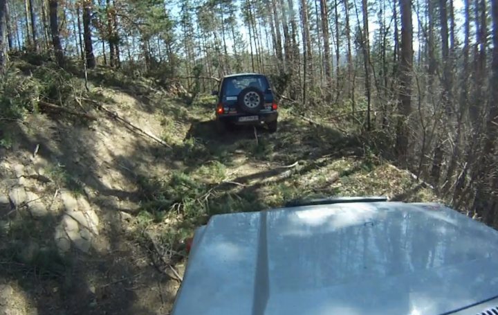 Bit of off-roading, and a bit of camping. - Page 1 - Off Road - PistonHeads - The image depicts an off-road scene where a vehicle is driving on a dirt path surrounded by overgrown vegetation. The vehicle, which appears to be a small pick-up truck, is on an incline and is heading towards a small wooded area. The terrain features a mix of rocky slopes and sparse plants, suggesting a wild or natural environment. Above, the sky is partially cloudy, hinting at the possibility of changing weather conditions. The perspective of the image suggests it was taken from another vehicle or a raised point, capturing the intrepid journey of the truck.