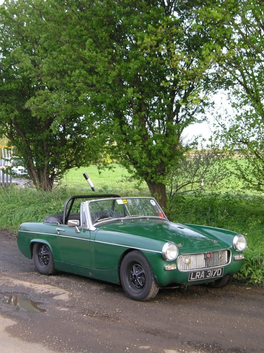 My Midget  - Page 1 - MG - PistonHeads - The image features a green vintage sports car parked on a dirt road. The car has a convertible top, which is currently up, providing a clean, sweeping appearance. It has round white headlights that contrast with the vehicle's green body. The background showcases a tranquil setting with trees, grass, and a field, emphasizing the contrast between the old-fashioned vehicle and the natural environment. The car exhibits some wear and age with minor scratches and dents, suggesting it has been driven and enjoyed despite its age.