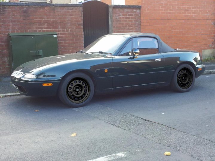 My BRG MK1 MX-5  - Page 1 - Mazda MX5/Eunos/Miata - PistonHeads - The image shows a dark-colored, potentially older-model, car parked on the side of a road. The car has a sloped front, a black beach-style saggy top car roof kit, and a distinctive design with wheel arches. There are no visible license plates, and no other vehicles or people are seen in the immediate vicinity. The road is smooth, and there is a cobblestone wall in the background to the left of the car.