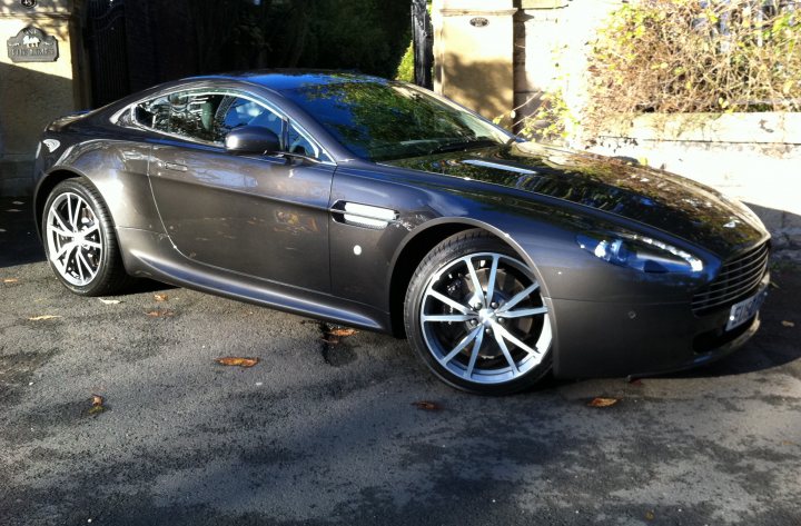 6 month update V8V - Page 1 - Aston Martin - PistonHeads - The image shows a sleek, dark-colored coupe parked on a driveway. The car has six-spoke wheels and is positioned with a prominent turn of the front wheel, suggesting the car is either entering or leaving the viewer's perspective. It appears to be a modern model, resting in an area with gravelly terrain. In the background, there are trees and foliage, with some fallen leaves scattered around, indicating an autumnal setting.