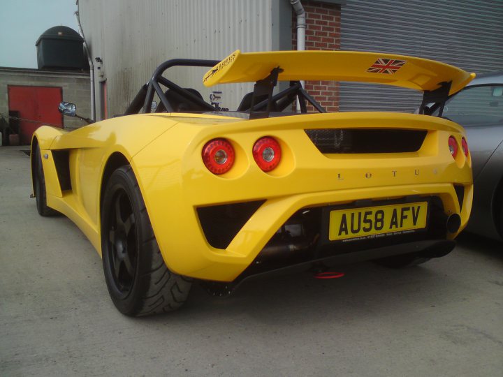 Pistonheads - This image features a sleek, yellow sports car that resembles a Lotus model. The car is parked on a concrete surface and it's facing towards the right side of the image. The bonnet of the car is adorned with a Union Jack flag, adding a distinctive touch to its appearance. Further down the right side, there is a glimpse of another vehicle, hinting at a shared parking space.