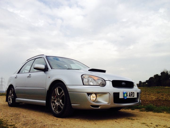 A white car parked in a parking lot - Pistonheads - The image shows a silver car parked on a gravel road with a plain, open sky and overcast weather. The car is angled towards the viewer, and there is a plate on the front with the letters "ARD" displayed. The car features a raised silver-colored spoiler and multi-spoke alloy wheels. In the clear background beyond the vehicle, a power line can be spotted, and the lighting suggests either dawn or dusk.