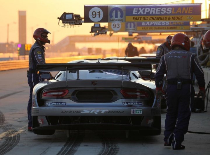 New SRT Advert - Page 1 - Vipers - PistonHeads - The image portrays a scene at what appears to be a Formula 1 pit stop. In the foreground, two individuals, likely crew members, are standing next to a silver and black race car, bending over it with a focused demeanor. They are both wearing red helmets and are dressed in crew attire with the word 'MICHELIN' visible on their outfits, suggesting they might be there to service the car's tires. In the background, there's a glowing sign overhead that reads 'EXPRESS LANE', indicating the location's name, and another sign that seems to be related to exhaust changes and more fast-paced topics. The setting is an outdoor race track at either sunrise or sunset, as indicated by the warm tones of the sky. The overall scene captures the intense and precise atmosphere of an F1 event.