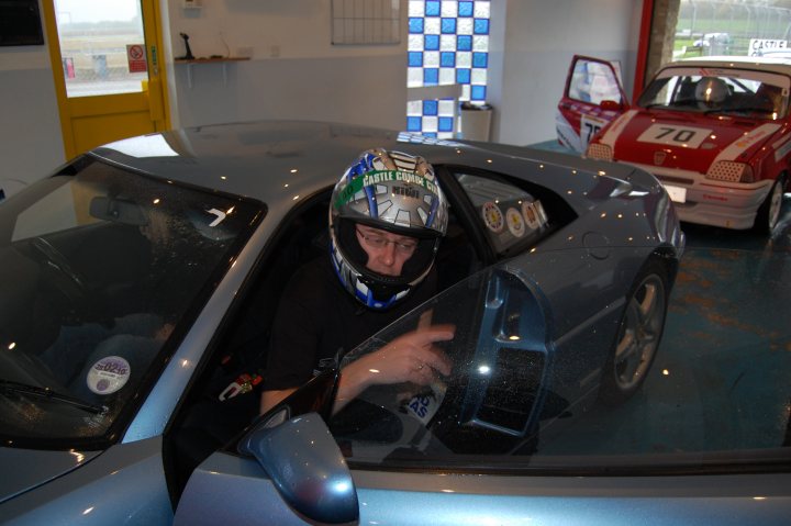 Association Stroke Combe Castle Supercar Pistonheads Day - This image portrays a scene inside a display of a car showroom. A man is positioned inside a silver sports car, which is open and facing him. He is wearing a gray helmet, indicating that he is likely a participant in the show. The car is equipped with a sunroof and is designed with aerodynamics in mind. There are other cars visible in the background, suggesting a variety of vehicles on display. The lighting in the room is bright, revealing reflective surfaces, and the overall atmosphere seems clean and polished.