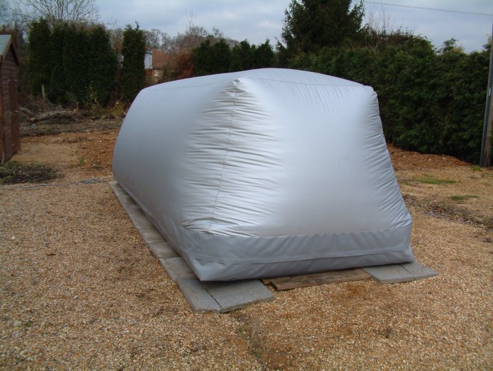 Carcoon or ???? Outdoor car storage - Page 1 - General Gassing - PistonHeads - The image captures an unusual scene: a large, inflatable structure, wrapped in a shiny, metallic foil, stands alone in a gravel driveway. The foil wraps tightly around the object, reflecting the light in a way that catches the eye. In the background, there's a hint of greenery, suggesting that the gravel driveway might be in a residential area or a garden. The surroundings are otherwise bare, emphasizing the size and unusual nature of the wrapped object. The scene's simplicity and stark foil contrast give it an almost sculptural quality.