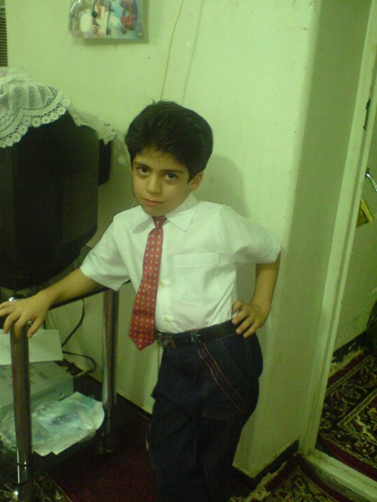 This is a candid photograph of a young boy with brown hair styled in a side parting. He is wearing a white dress shirt with a red tie and darker pants that appear to be jeans. The boy has a slightly puzzled or uncertain expression and is posing with his left hand resting on his hip. The room in the background is somewhat messy, with objects such as books and a stack of paper on a table, among other items. The setting suggests a personal space, potentially a home or a family room.