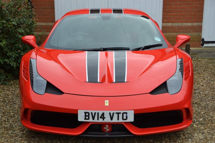 458 Speciale - Page 1 - East Anglia - PistonHeads - The image shows a red sports car that appears to be resting on a cobbled or gravel driveway. The car is a model by Ferrari, as indicated by the yellow and black emblem on the hood and the logotype on the license plate. The license plate itself reads "BV14 VTO." The vehicle has a sleek, aerodynamic design typical of high-performance cars, and its side windows reflect the environment. The headlights and side mirrors are distinctive, suggesting a high-end model, and the overall impression is that of a luxury, high-performance sports vehicle.