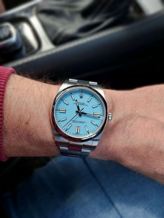 Wrist check 2021 - Page 60 - Watches - PistonHeads UK - The image shows a person wearing a watch on their left wrist. The watch is blue and has a silver band, featuring a red second hand. It appears to be a Rolex model. The person is sitting in the driver's seat of a vehicle, as indicated by the steering wheel visible in the background. The individual is dressed casually with a red long-sleeved top. The setting suggests a calm interior environment. There are no visible texts or distinguishing brands within the image.