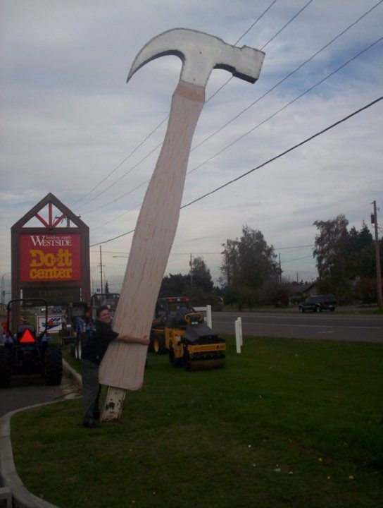 Pistonheads - The image captures an outdoors scene where a man is playfully posing with oversized construction equipment, including a giant hammer and a long piece of timber, which are humorously positioned to simulate being pulled out of the ground. The setting appears to be near the Westside Hardware Store, as indicated by a store sign in the background and various hardware displays. The environment suggests a street with grass and shrubbery, and there are mentioned flying monkeys and benches, but there is no sign of these in the visible portion of the image. The presence of other construction equipment and vehicles nearby adds to the whimsical and exaggerated nature of the scene.