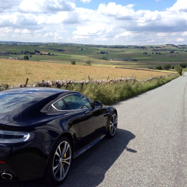 A car is parked on the side of the road - Pistonheads - The image showcases a sleek black sports car parked on the side of a winding country road. The road is flanked by a sturdy stone fence, beyond which vast expanses of open fields stretch into the distance, interspersed with distant hills. The sky is a serene blue, speckled with white clouds that add to the tranquil rural scene. The car's design and the pristine natural setting create a striking contrast, suggesting a sense of adventure or luxury escape to nature.