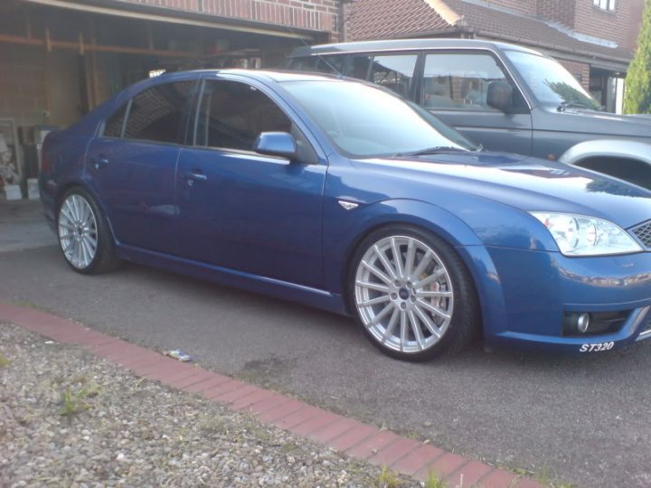 Upgrade Pistonheads Mondeo Questions - The image shows a vibrant blue sedan parked on a street alongside a brick sidewalk. The vehicle's sleek design and alloy wheels suggest it might be a newer model. Behind the sedan, there is a hint of another vehicle, which is too small to identify clearly. The setting appears to be residential, with a house visible in the background. The overall ambiance of the image suggests a calm and quiet suburban area.