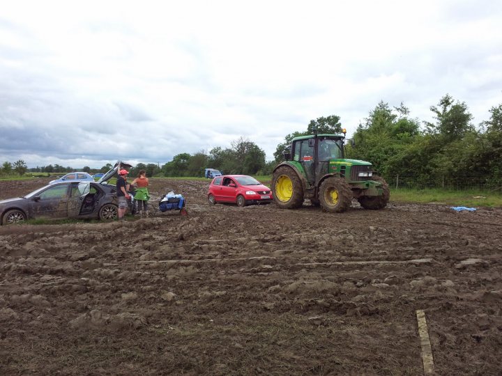 Silverstone F1 parking or park & ride? - Page 1 - Formula 1 - PistonHeads - In the midst of a muddy field, a green and yellow agricultural tractor becomes the center of attention. The farmer's family stands near their dirty red compact car, both their car and the tractor having been used extensively in recent muddy field activities, as evidenced by the mud splattered all over them. An orange car, perhaps a four-wheeler, is parked nearby, adding to the off-road ambiance. A wooden fence encloses the scene, indicating the boundary of the agricultural field. The atmosphere is realistic, painted with hues of earthy tones and the clear sky above, suggesting a typical day on the farm for the family.