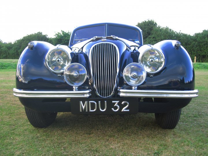 Fixed Pistonheads Head Coupe - The image shows a classic, convertible vehicle parked in a grassy field. The car is an antique model, exhibiting a deep blue color with chrome accents on the grille and the headlights. The headlights are round and prominently feature the classic diamond pattern. At the front of the car, the license plate reads "MDU 32," indicating it may be registered or used in Denmark. The background is blurred but suggests a natural, outdoor setting with trees at the far end of the field.