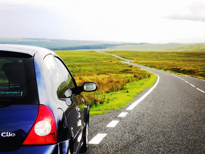 Pistonheads French Rattly - The image shows the rear end of a blue car with the Clio badge prominently displayed above the brake light. The car is stationary on a paved road, characterized by a white line on each side. The surrounding scenery features lush green hills or valleys that stretch into the distance, culminating under a sky of white. The overall impression is that of a peaceful road trip with clear weather conditions.