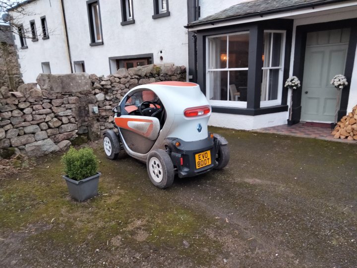 Hardcore EV... - Page 1 - EV and Alternative Fuels - PistonHeads UK - The image features a small, two-wheeled vehicle parked on a gravel driveway. This vehicle has a unique design with a white and orange color scheme, and it appears to be an electric or hybrid model, as indicated by the visible charging port at the rear. The setting is a residential area, characterized by a stone house in the background, with a neatly trimmed lawn and a well-maintained driveway. The overall atmosphere of the image suggests a peaceful, suburban environment.