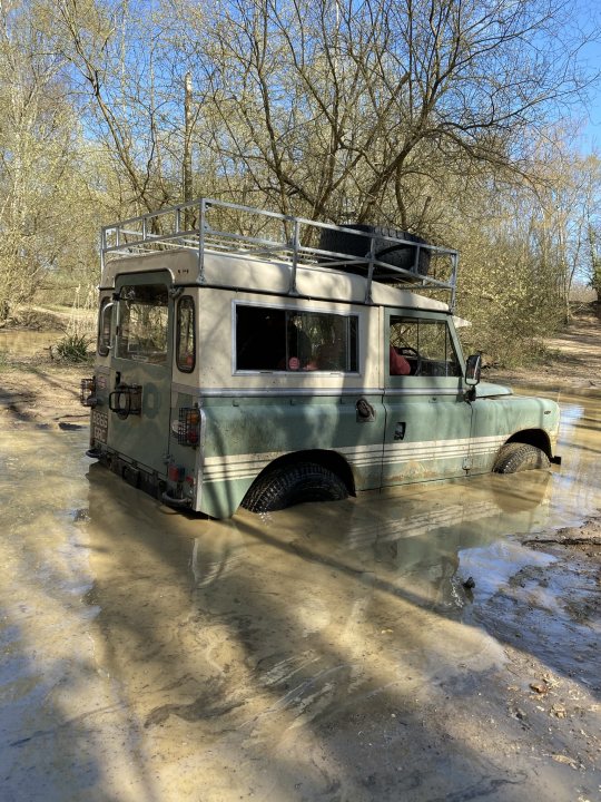 Pictures of your Classic in Action - Page 23 - Classic Cars and Yesterday's Heroes - PistonHeads UK - The image depicts a scene in nature where a vintage truck is driving through a muddy path. The truck, with its open sides and tall roof rack, stands out amidst the greenery surrounding it. It appears to be navigating through a wooded area, as suggested by the trees visible in the background. The environment seems tranquil, yet challenging due to the muddy conditions of the road.