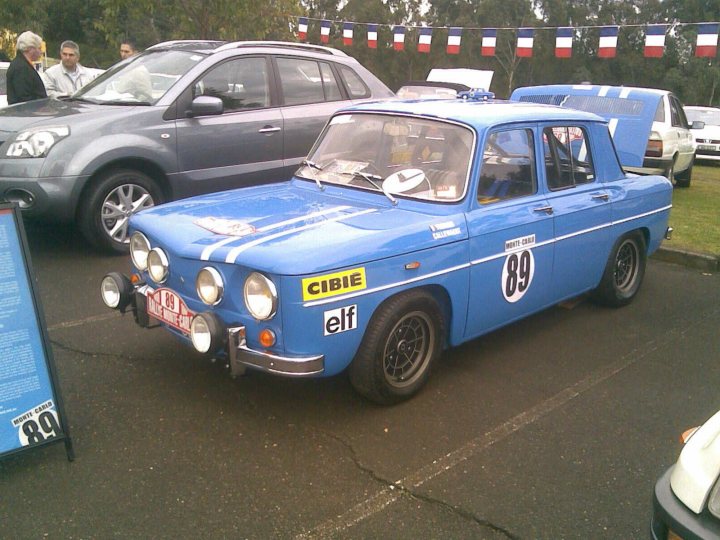 Pistonheads Add Renault - The image captures a scene of a small blue car, parked in a lot where various other cars are also visible. The blue car is adorned with yellow-framed headlights and sports a stripe on its hood. There's a sign on the windshield with a car emblem and the number '83'. A black and white striped awning towers above the car, suggesting the presence of a nearby business or event. In the background, a tall flag pole with multiple colorful flags can be seen, adding to the visual interest of the scene.