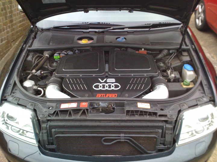 Dyno Mrc Pistonheads - This image depicts the engine bay of a car, which is open, revealing the car's V8 engine. The engine has a dominant presence, with its exposed components, such as the metal parts, the intake manifold in the center, and several ribbed panels. There are several emblems and decals on the engine, and two coolant bottles are placed in front of it. The car trunk is open above the engine, revealing a dark interior. Another car is visible in the background.