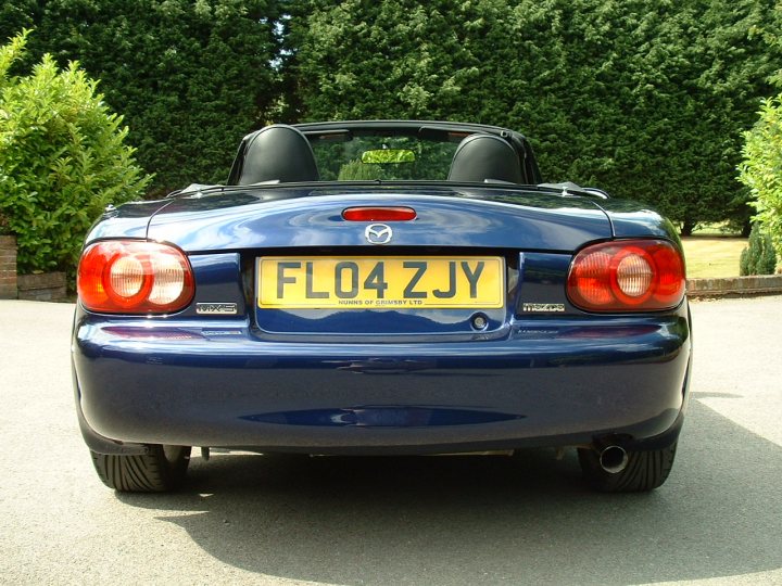 Pistonheads Stopping - This image shows a dark blue Mazda Miata sports car parked outdoors. The car is viewed from a rear angle, highlighting the license plate, which reads "F1O4 Z3JY," and the right tail-light. The surrounding area includes a tarmac driveway and green hedges, suggesting a residential setting. The sky is visible behind the car, providing natural light for the scene.