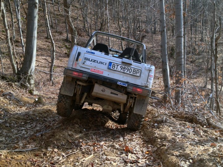 Bit of off-roading, and a bit of camping. - Page 1 - Off Road - PistonHeads - In the image, you can see a Suzuki Bt990 BP off-road vehicle in an off-road setting. The 4x4 vehicle is navigating a wooded area with small trees and leafy ground, indicating that it is possibly on a rugged course. The vehicle's condition appears to be well-maintained, and it has a small sticker on the back. The image captures the vehicle tilted to one side, suggesting that it might be driving on an incline or negotiating a challenging section of the track. The surroundings of wood and grass are indicative of a natural environment, such as a mountain or forest trail.