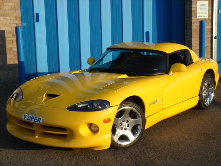 Hello! - Page 1 - Vipers - PistonHeads - The image shows a striking yellow sports car parked outside a building with blue doors. The car's design appears modern and it occupies most of the frame. It features side scoops, large alloy wheels, and a sleek body that suggests high performance capabilities. The car is parked on an asphalt surface, and the overall mood of the image is bright and energetic due to the vibrant yellow color of the car and the clear daylight.